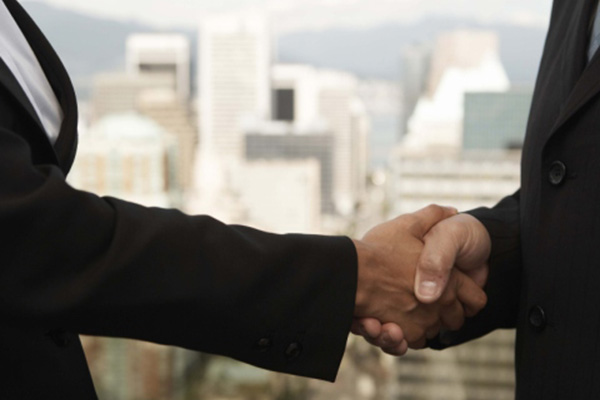 Businessman and businesswoman both wearing black suits and shaking hands with city visible in background