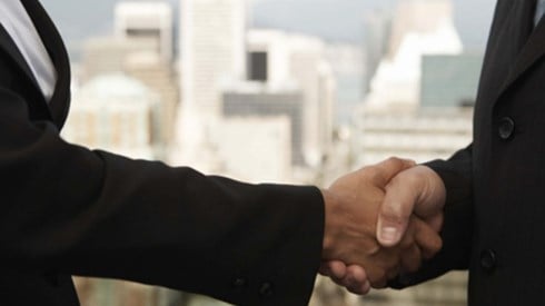 Businessman and businesswoman both wearing black suits and shaking hands with city visible in background