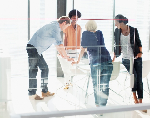 A group of 4 millennials standing around all four sides of a drafting type table in a glass office workplace