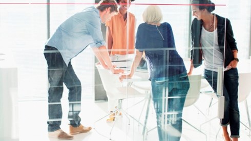 A group of 4 millennials standing around all four sides of a drafting type table in a glass office workplace