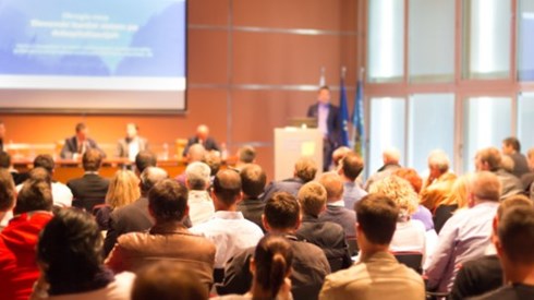 Group of people viewing a slide presentation in a large room with speaker at podium, panel of experts, and 3 flags in corner