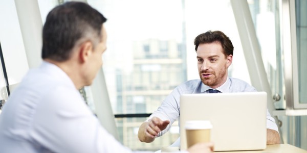Two businessmen in a meeting with one using a laptop and the other listening with a coffee cup