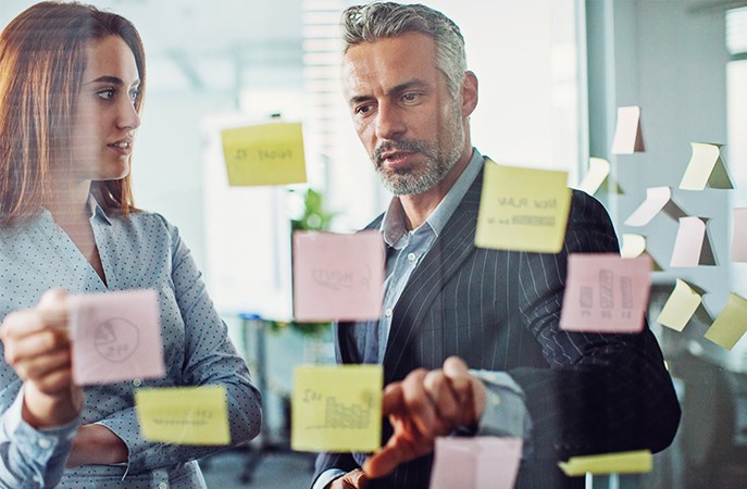 Business colleagues, man and woman, discussing data comparison on postit notes on glass wall