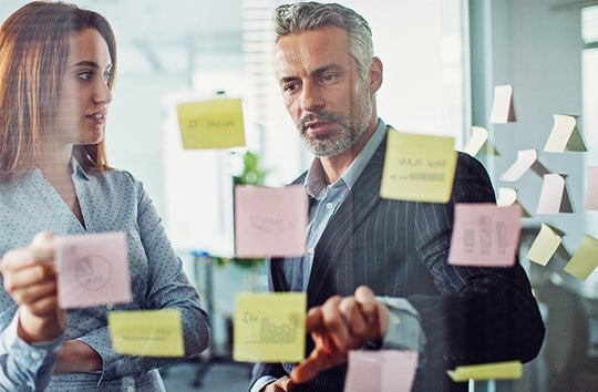 Business colleagues, man and woman, discussing data comparison on postit notes on glass wall