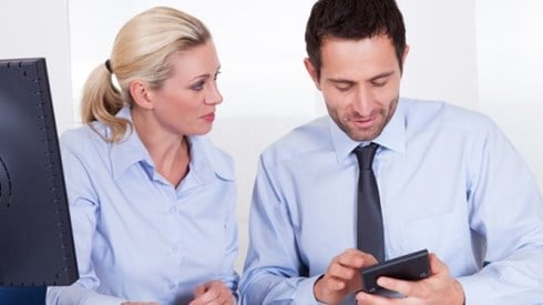 Woman looking at man using a hand held device in an office setting in front of a monitor