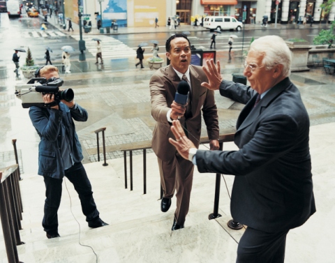 An older man in a blue suit holding up his hands against approaching reporter and cameraman on steps of a building in a city