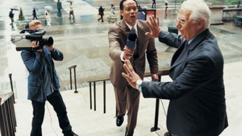 An older man in a blue suit holding up his hands against approaching reporter and cameraman on steps of a building in a city