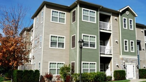 Green and taupe apartment complex building with green grass, a lamp post, and a tree with orange autumn leaves