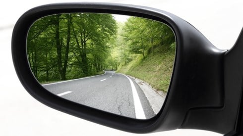 Vechicle rearview mirror showing road with green trees in background