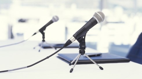 2 tabletop microphones sitting in front of black notebooks with a pen on top at a white table with chair