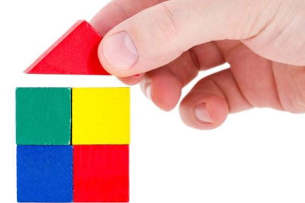 Hand placing triangular red building block on four stacked red, yellow, green, and blue blocks in the shape of a house