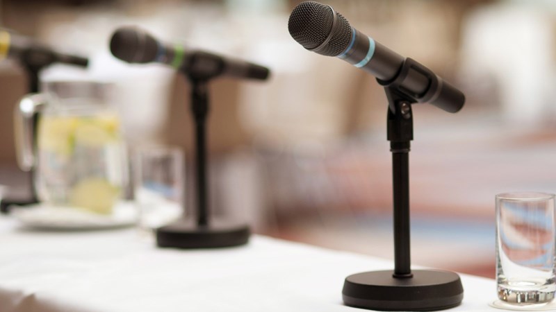 Three black microphones on a white table with a pitcher and glass of water
