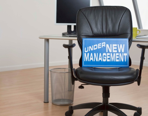 Blue sign with the words UNDER NEW MANAGEMENT sitting on a black leather chair in an empty office