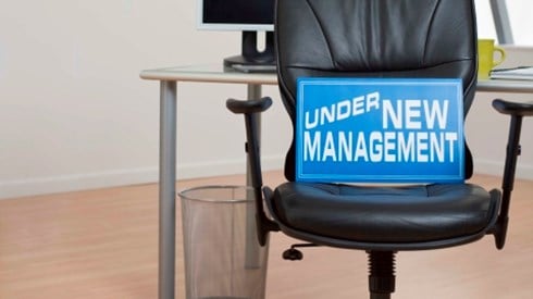 Blue sign with the words UNDER NEW MANAGEMENT sitting on a black leather chair in an empty office