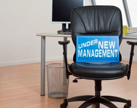Blue sign with the words UNDER NEW MANAGEMENT sitting on a black leather chair in an empty office