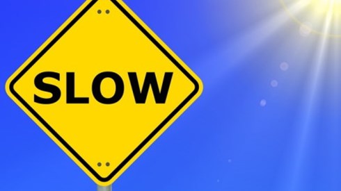 Yellow diamond-shaped traffic warning sign that says SLOW and clear blue sky and partial sun and rays in background