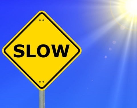 Yellow diamond-shaped traffic warning sign that says SLOW and clear blue sky and partial sun and rays in background