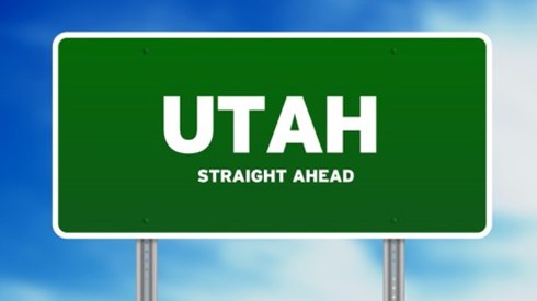 A green road sign in front of a cloudy blue sky with the words Utah and straight ahead written on it.
