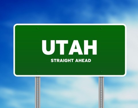 A green road sign in front of a cloudy blue sky with the words Utah and straight ahead written on it.