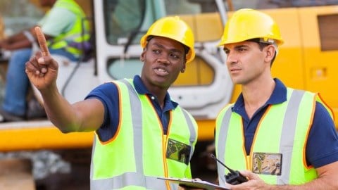 Two construction workers at a site discuss the project and another is operating a large construction vehicle in background