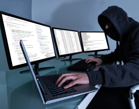 A hooded computer man in front of 3 large monitors with one hand working from a laptop and the other from a computer keyboard