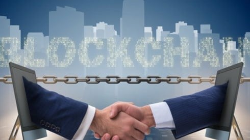 Two businessmen´s hands extend from two monitors to shake hands in front of a chain, the word Blockchain, and a city skyline