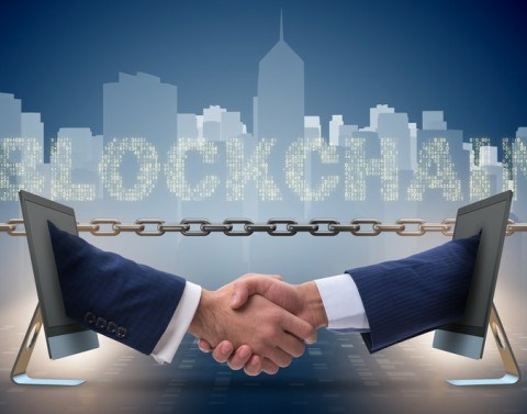 Two businessmen´s hands extend from two monitors to shake hands in front of a chain, the word Blockchain, and a city skyline