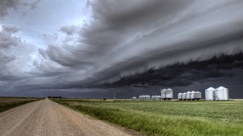 Large storm clouds hover over the flat land of a farm with silos and a dirt road separating crops.