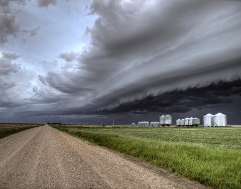Large storm clouds hover over the flat land of a farm with silos and a dirt road separating crops.