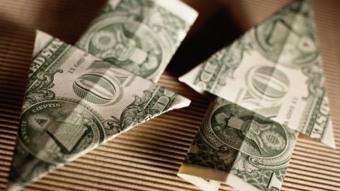 Two one dollar bills folded in the shape of arrows pointing in opposite directions sitting on brown corregated cardboard