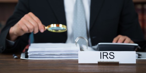 IRS desk sign and man in suit holding a magnifying glass over binder of documents while running a desk calculator