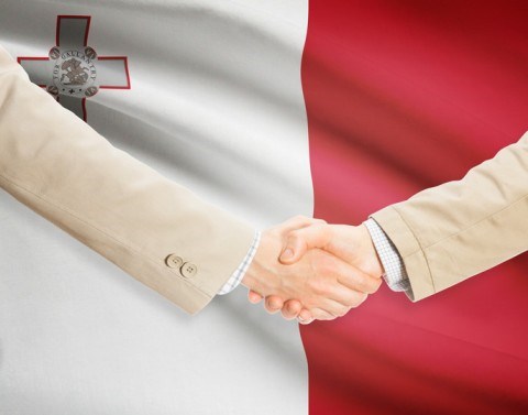 Two businessmen shake hands in front of the flag of Malta