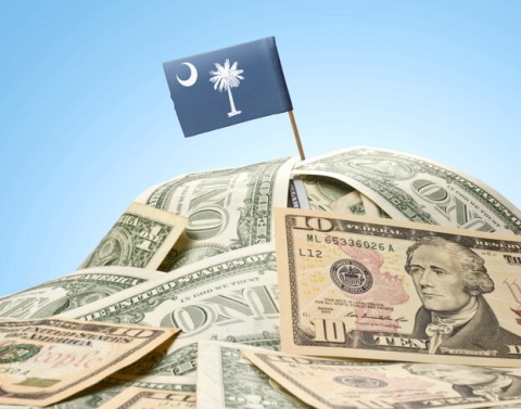 A hill-like stack of 1- and 10-dollar bills with a tiny flag pole and the South Carolina State Flag