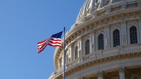 Close up of top portion of Capitol building with United States flag flying on a pole and blue sky in background