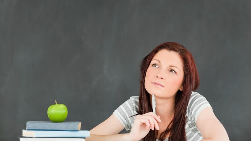 A student thinking and sitting at a desk that has an apple on top of a stack of books alongside other groups of papers