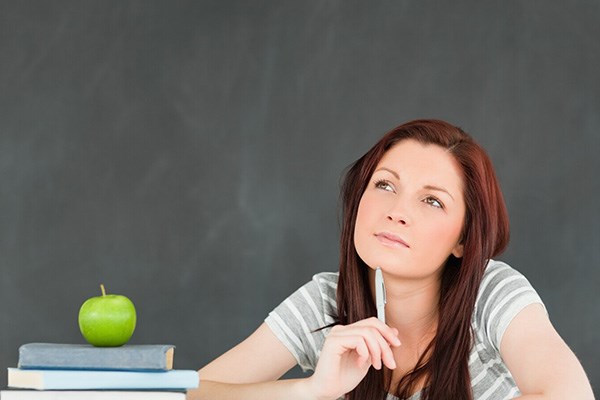 A student thinking and sitting at a desk that has an apple on top of a stack of books alongside other groups of papers