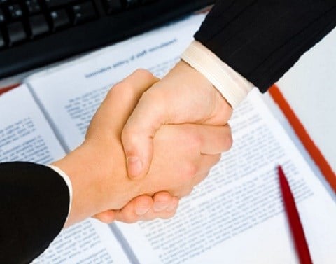 Two businessmen shaking hands over a contract in a red binder with a red pen and keyboard next to it