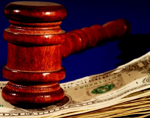A shiny all wood mahogany colored gavel resting on top of a pile of one dollar bills with a blue background