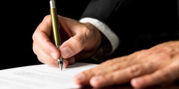 A businessman is using one hand to support a document and the other to sign it.