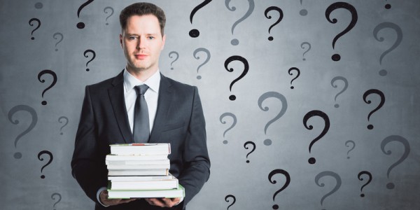 Businessman holding large bound references is surrounded by several black and grey question marks