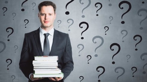 Businessman holding large bound references is surrounded by several black and grey question marks