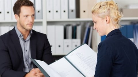 Woman holding a resume and interviewing a man sitting in an office with binders on shelves in the background