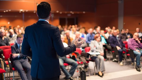 Man viewed from behind delivering speech at conference with crowd out of focus in front of him