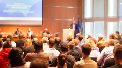 Group of people viewing a slide presentation in a large room with speaker at podium, panel of experts, and 3 flags in corner