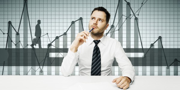 A young businessman sitting musing with a background that has bar and line graphs and a silhouette of a businessman walking