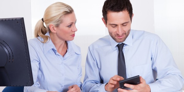 Woman looking at man using a hand held device in an office setting in front of a monitor
